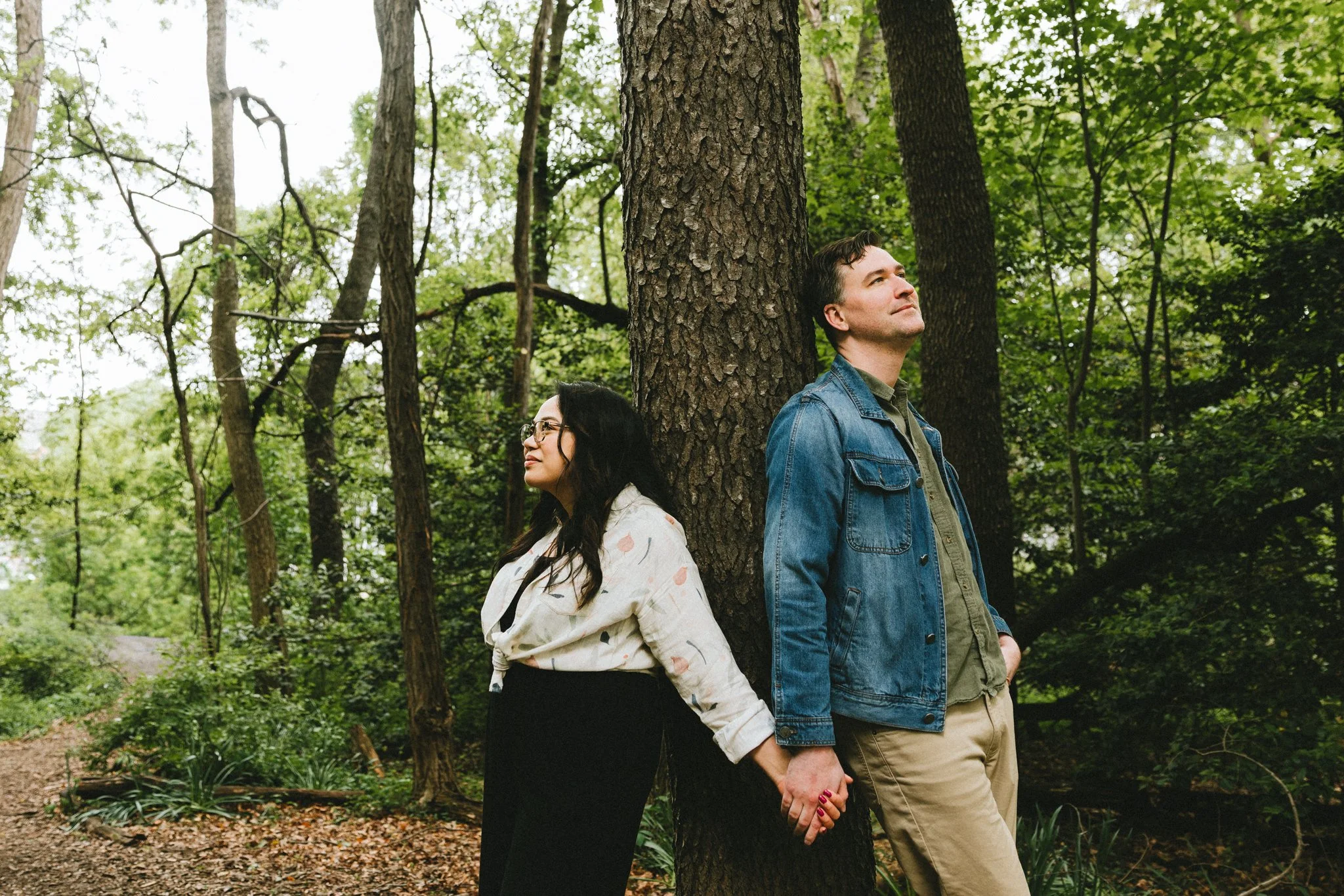  The engaged couple leaning against a tree trunk side by side, looking in opposite directions, holding hands 