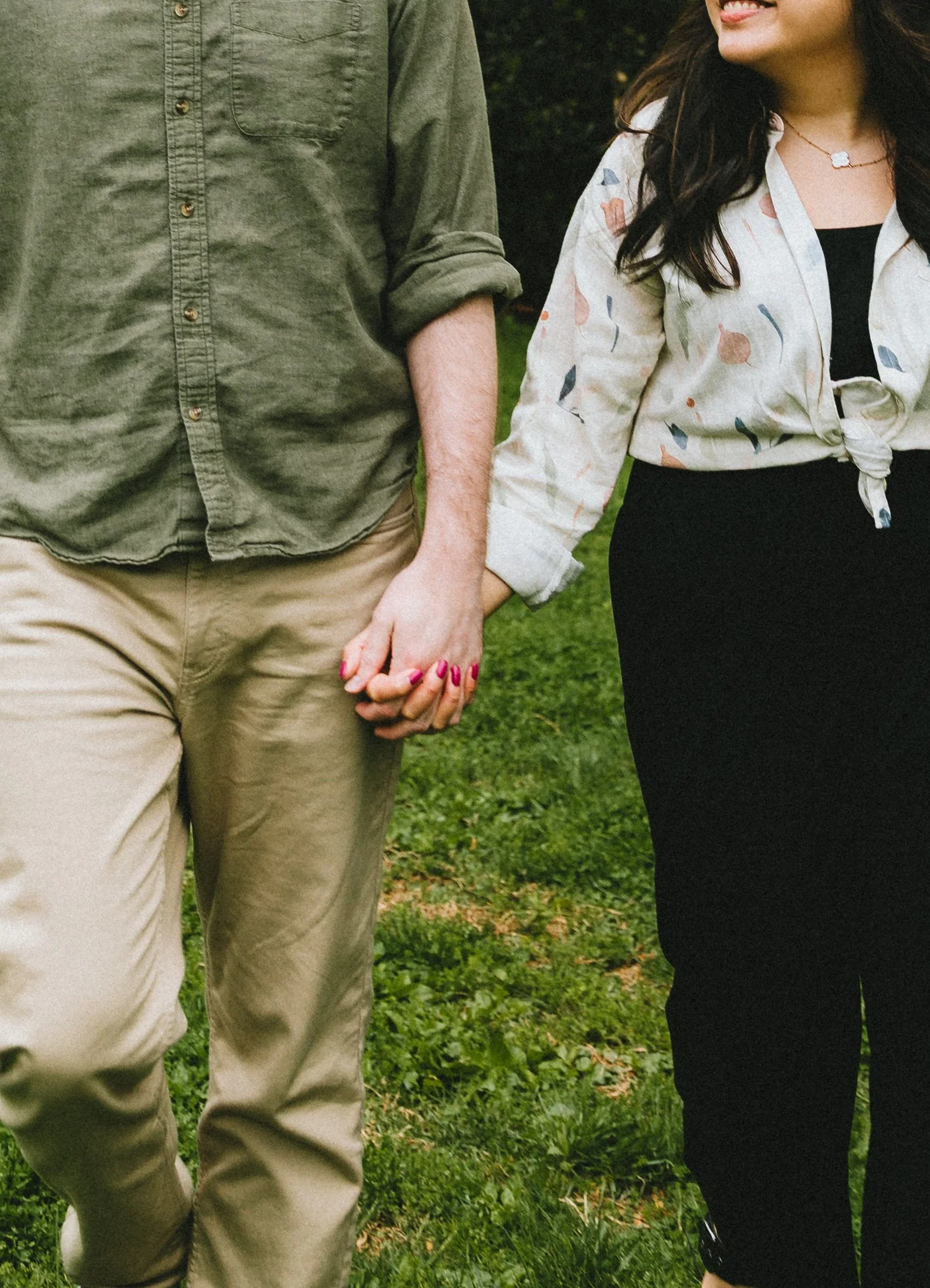  Close-up of the couple holding hands while walking on the grass, Mariel's necklace and patterned blouse visible 
