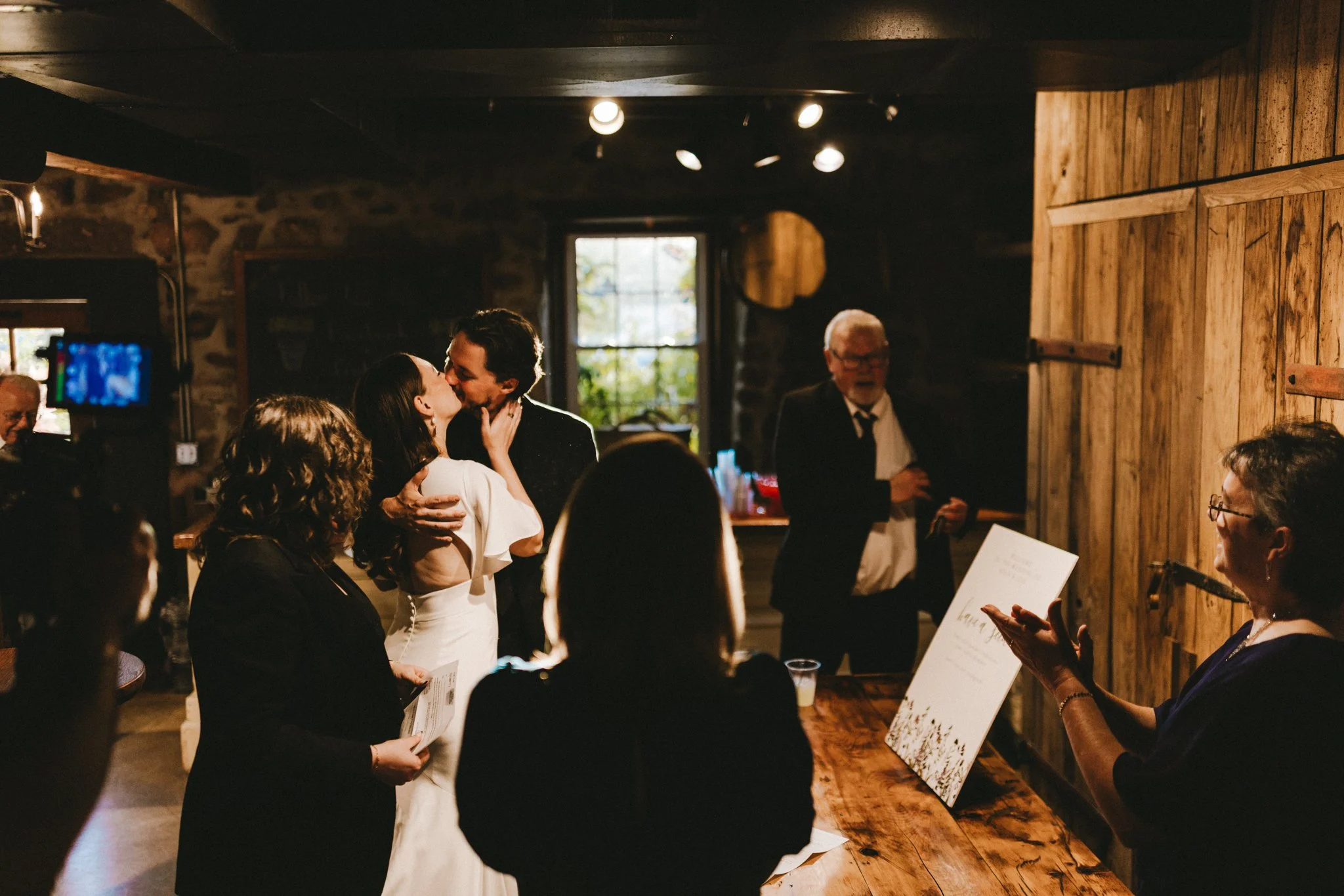 Bride and groom sharing a kiss inside a warmly lit barn venue, guests watching in the background