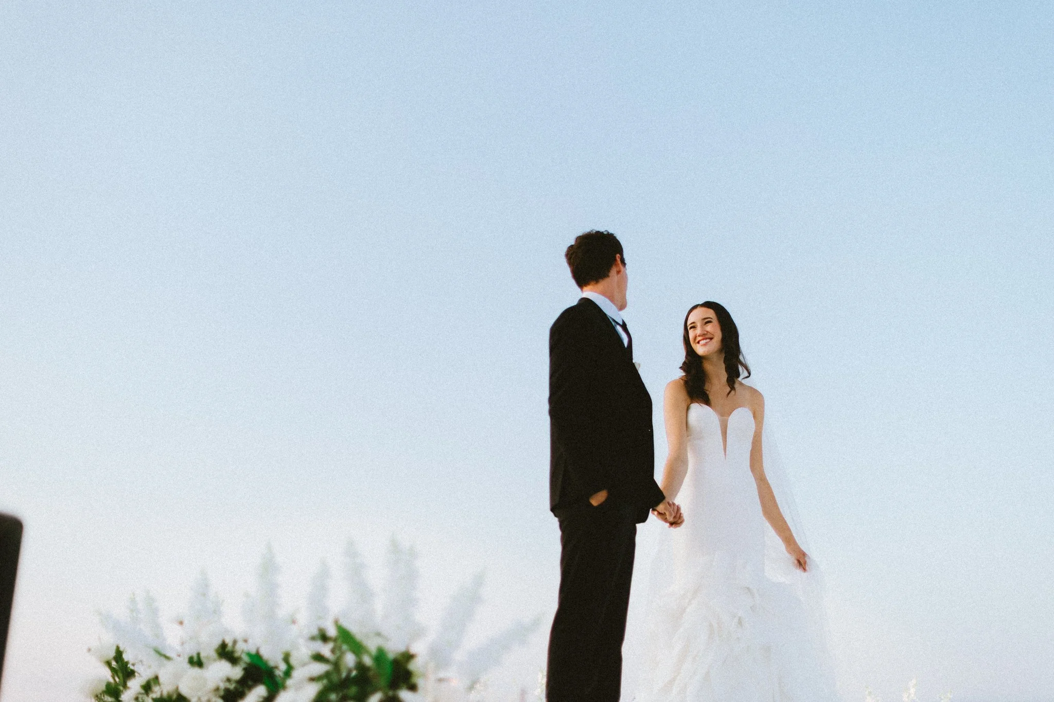 Bride smiling at groom while holding hands during outdoor wedding ceremony against blue sky, Love Story By Aira Maryland