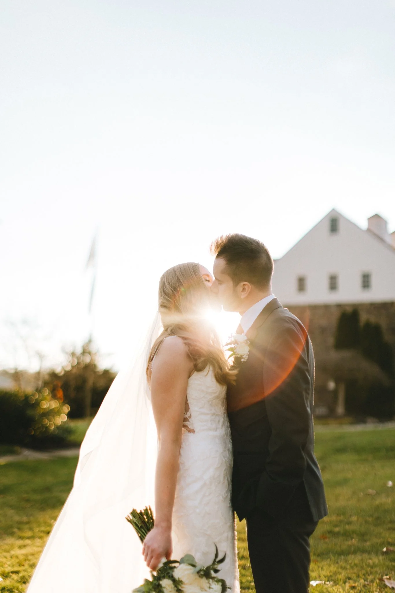 Bride and groom kissing at golden hour, sun flare breaking through between them, white farmhouse visible in the background