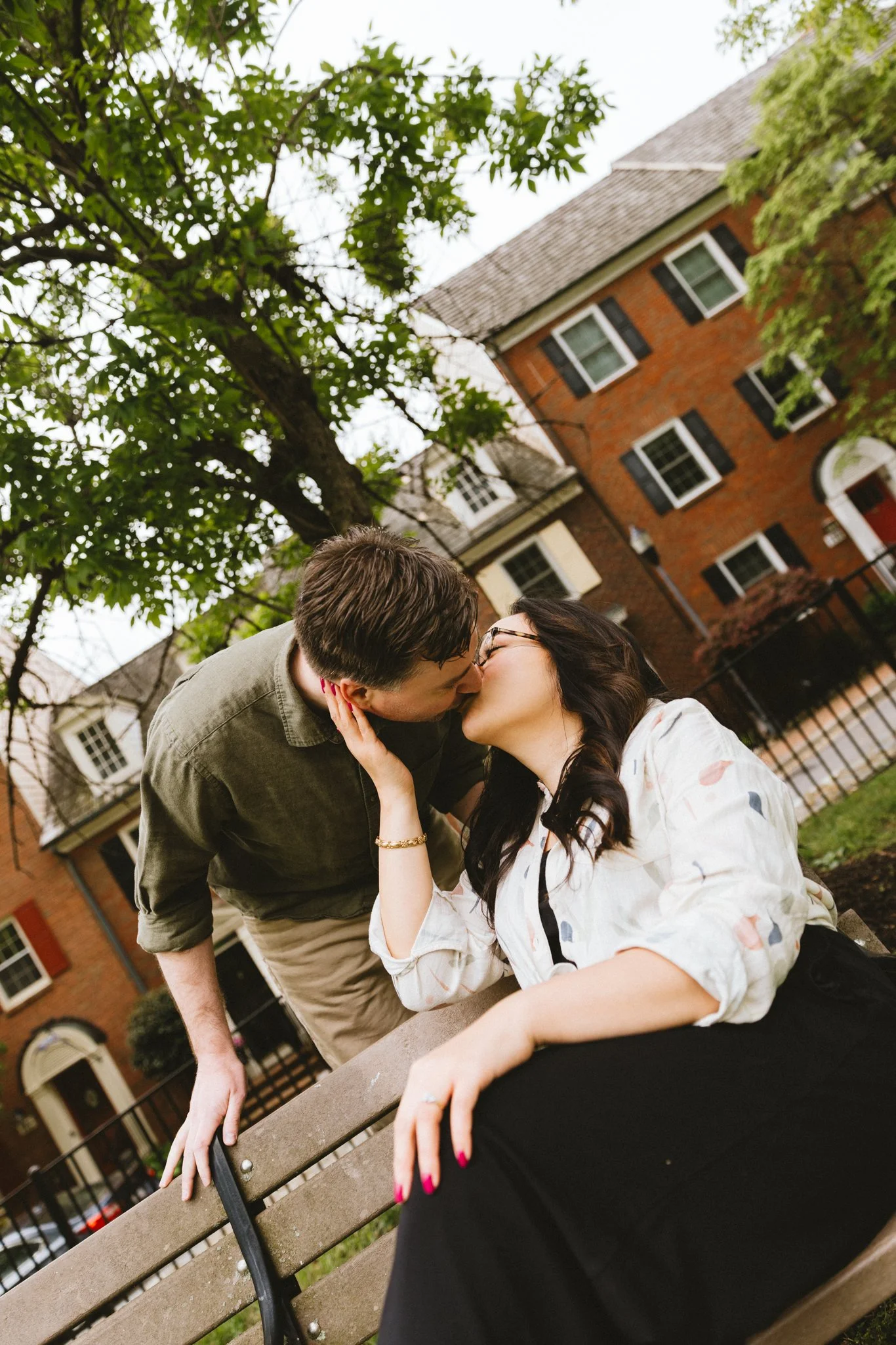  Ryan kissing Mariel from above as she leans back on the bench, brick townhomes with a red door in the background 