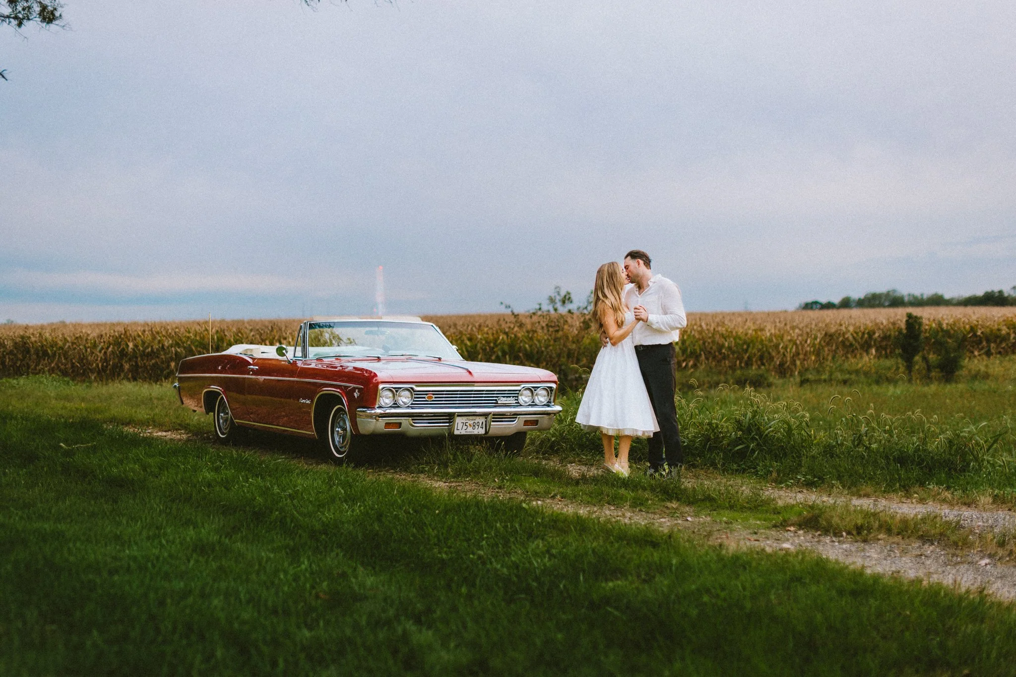 Couple sharing a romantic embrace beside a vintage red car in an open field, documentary wedding photography by Love Story By Aira Maryland