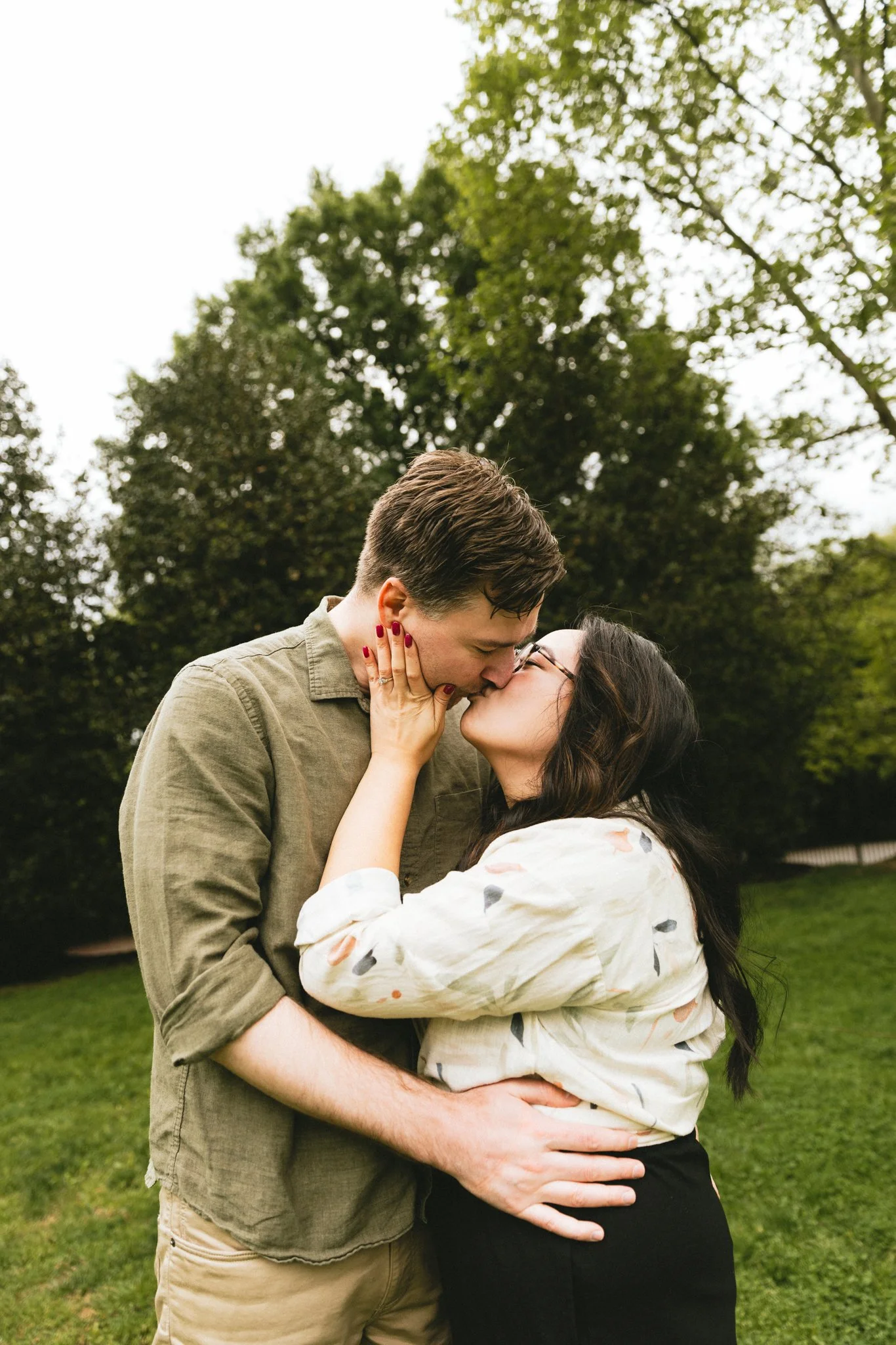  Mariel and Ryan kissing on the open green, Mariel's hand on his face, spring trees behind them — Love Story By Aira Photography 