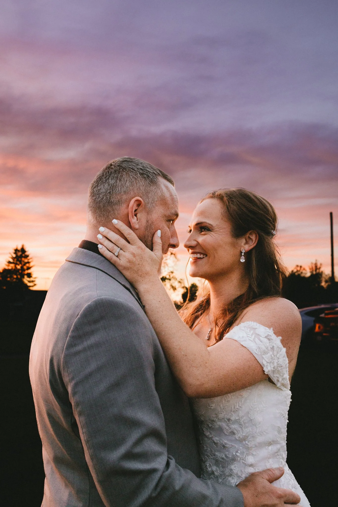 Bride holding groom's face as they lean close, the sky behind them deep purple and pink at blue hour