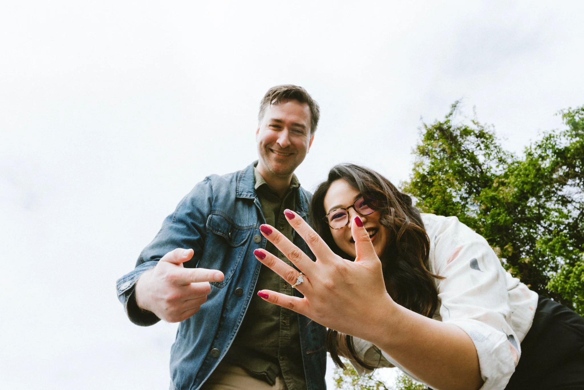  Mariel and Ryan both leaning toward the camera laughing, Mariel showing off her engagement ring — candid engagement session at Kentlands Green by Love Story By Aira Photography 