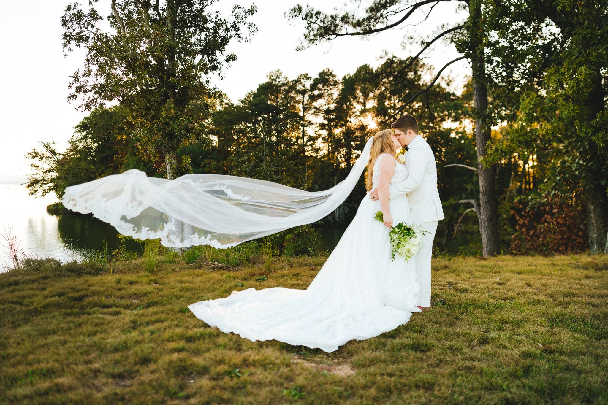 Bride and groom kissing as her veil flies in the wind, warm golden light breaking through the trees at Jubilee Farm in Maryland