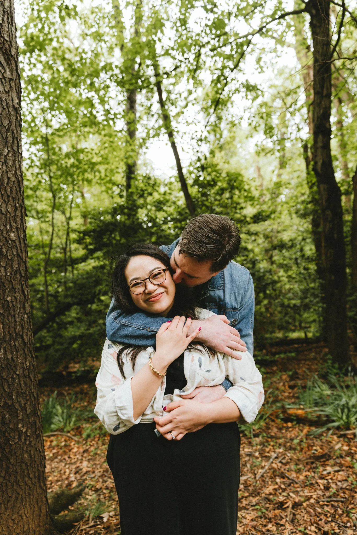  Ryan holding Mariel from behind, kissing her temple as she smiles with her eyes closed, surrounded by spring greenery 