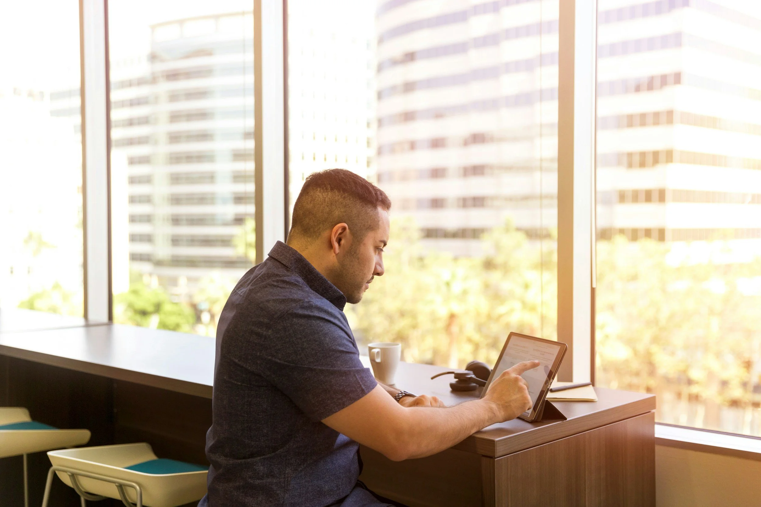 A man sitting at a desk near large windows using a tablet in a modern office building with tall glass skyscrapers outside.