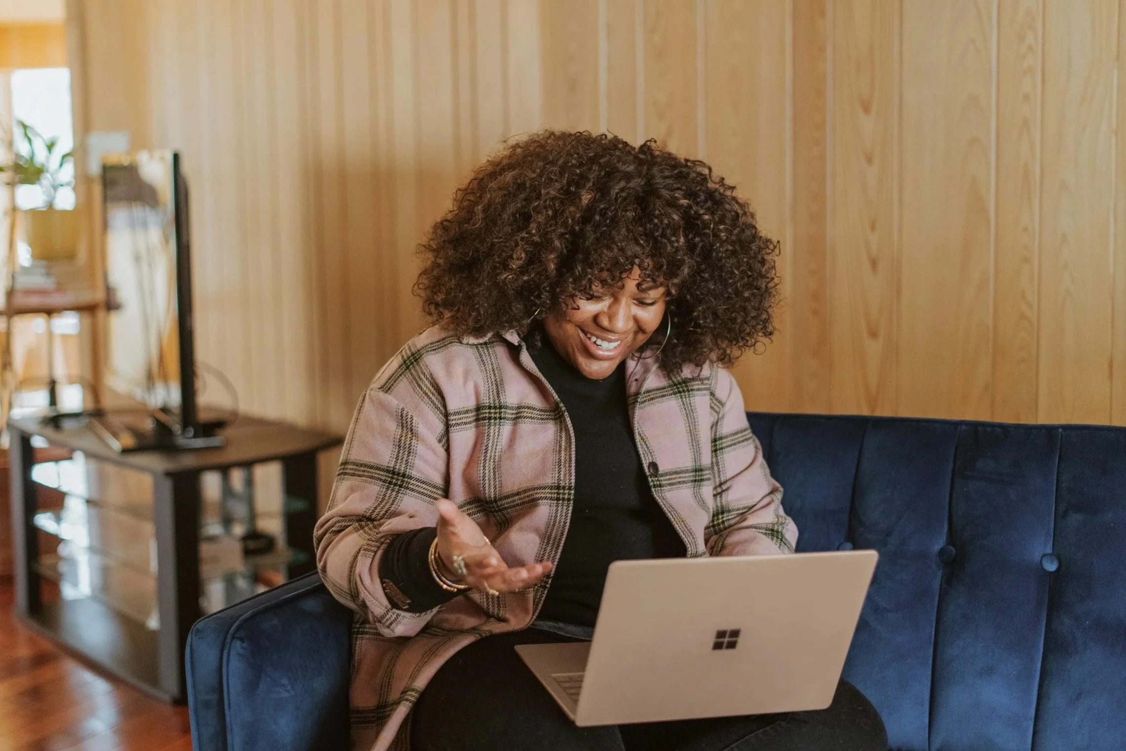 Woman with curly hair smiling while using a laptop in a cozy room with wood-paneled walls.