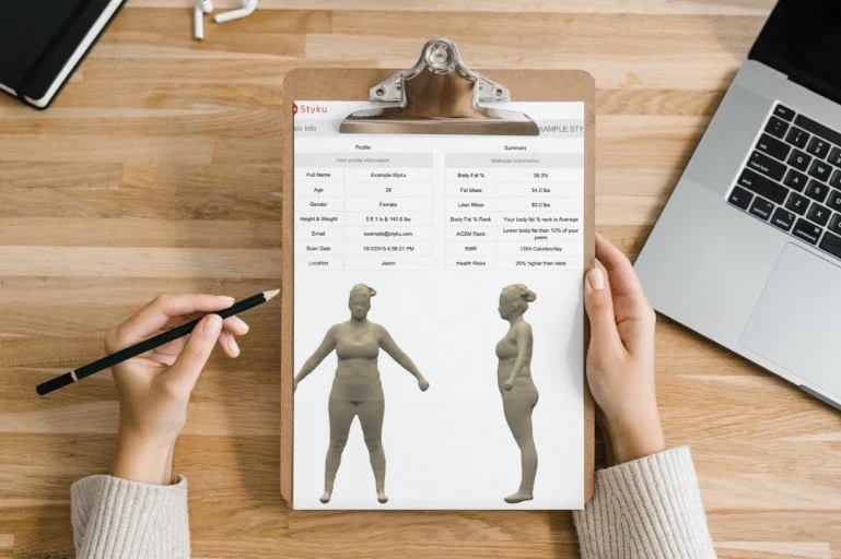 A person holding a clipboard with a body scan report displaying 3D models and information about body composition and measurements, next to a laptop on a wooden desk.