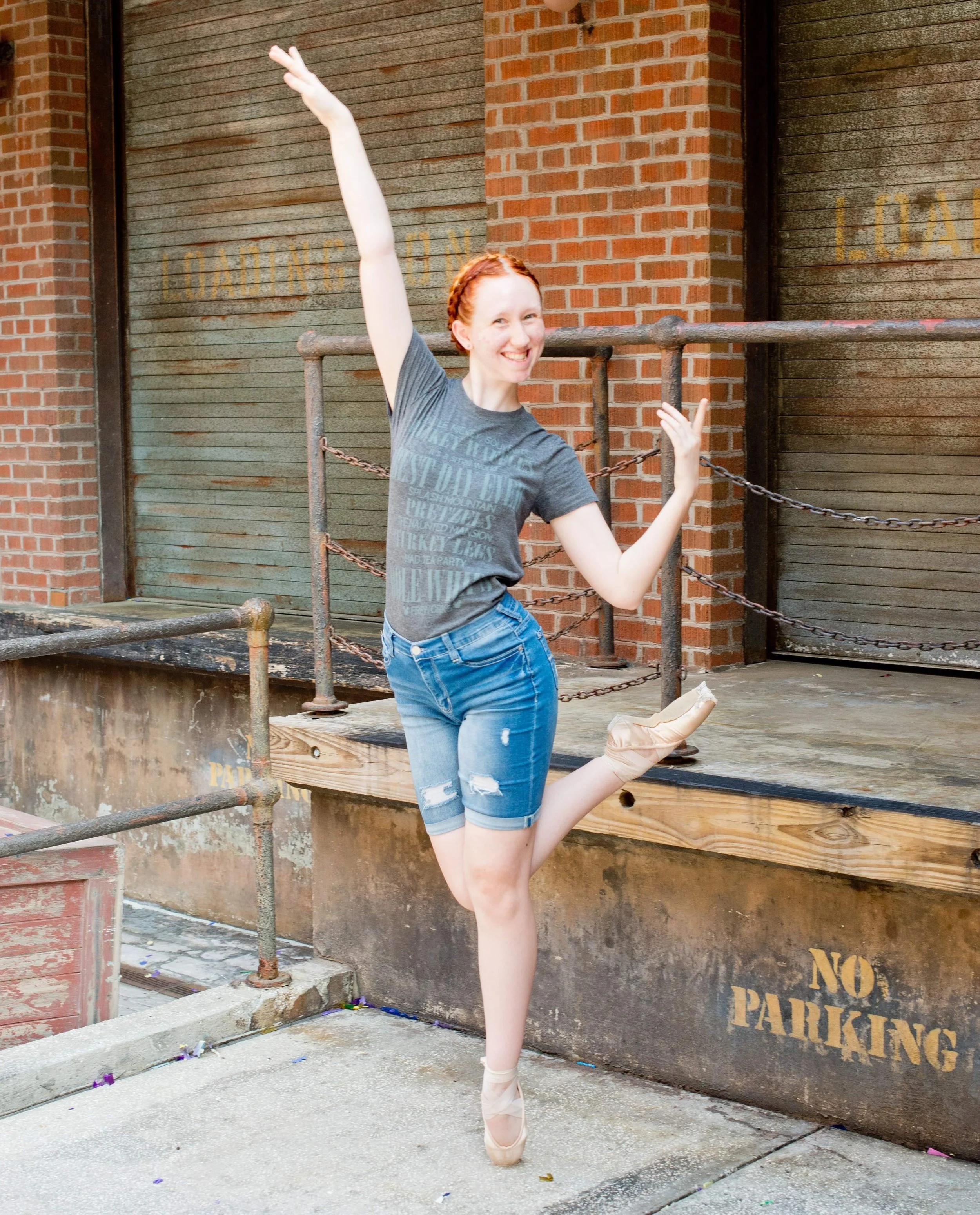 A ballerina with red hair in a gray t-shirt and denim shorts practicing ballet on pointe outdoors, standing on one foot with arms raised, next to a brick wall and a metal railing.