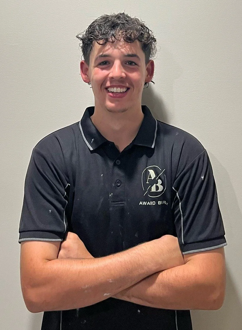 Young man with curly hair smiling, wearing a dark polo shirt with a logo on the chest, standing against a plain wall.