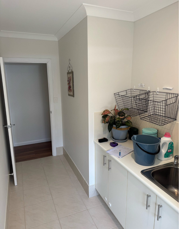 A small kitchen area with a white countertop, a sink, a potted plant, cleaning supplies, and hanging wire baskets. There is a doorway leading to another room with a wooden floor.