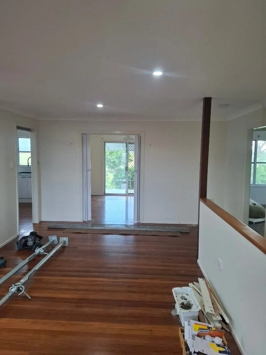 Interior of a house with wooden flooring, looking into a living room with sliding glass doors leading outside, some construction tools and materials on the floor.