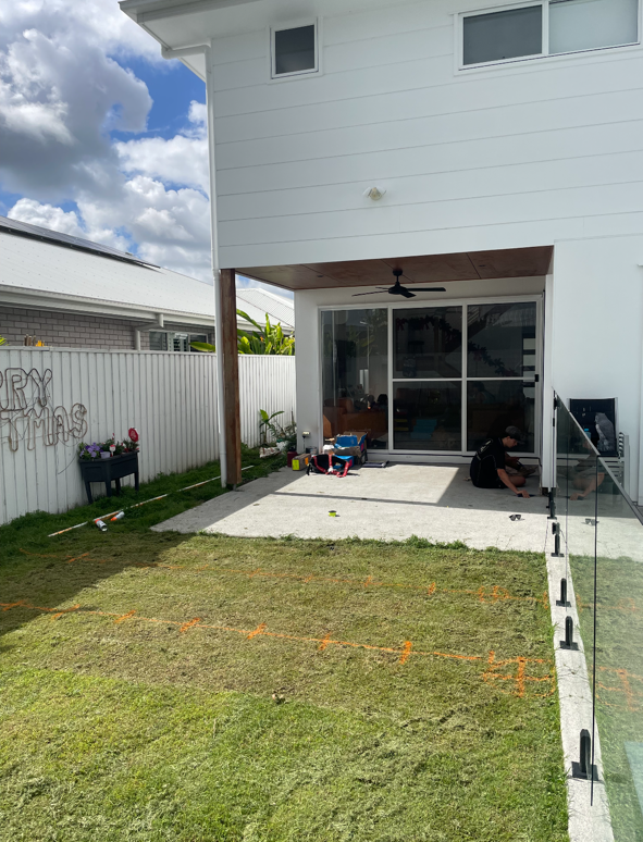 Backyard patio with a sliding glass door, a person sitting on the ground, tools and supplies, a white house with a balcony, and a fenced yard with a message on the fence reading 'Merry Christmas'.