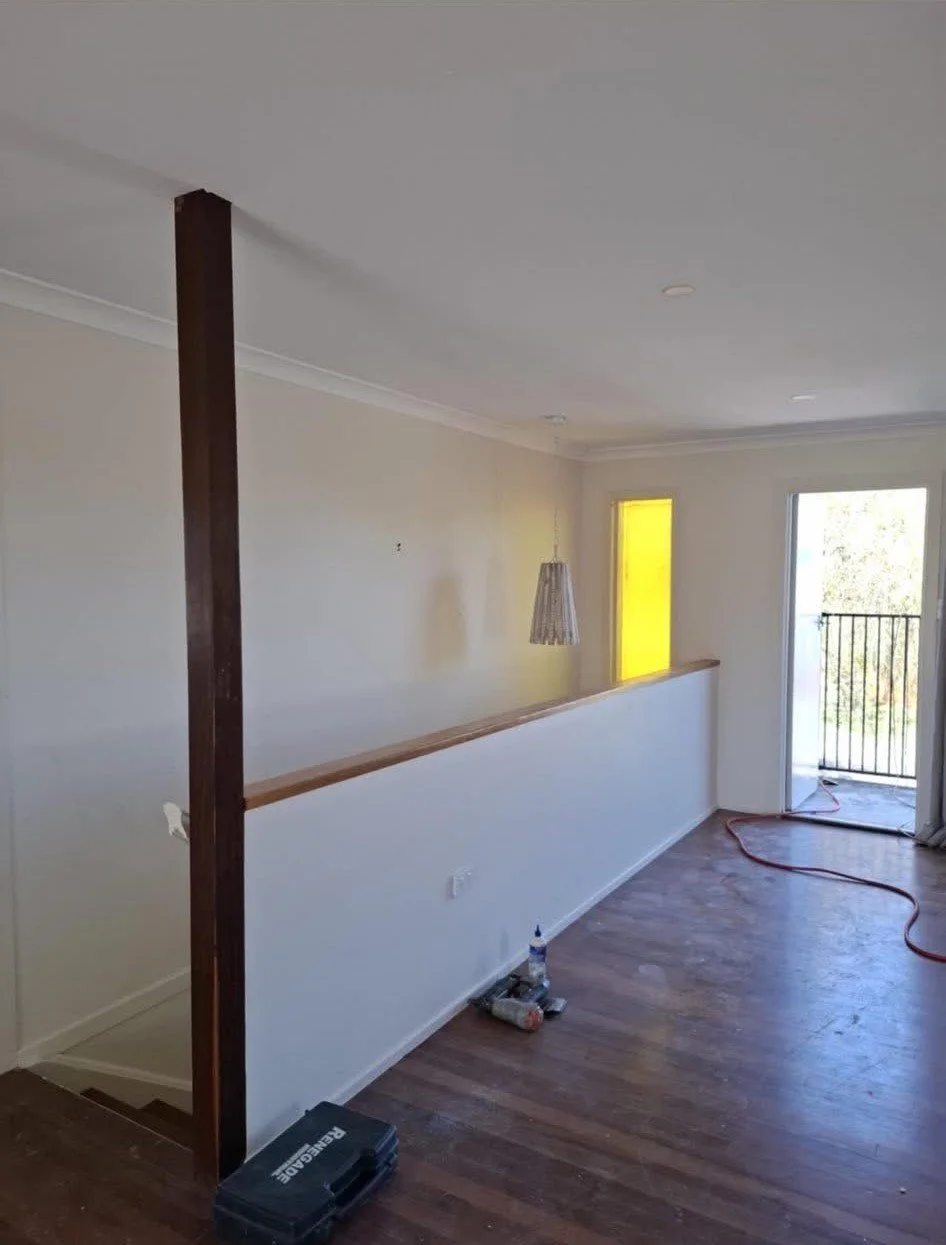 Interior of a room with a partially installed wooden railing, a hanging light fixture, and a balcony door. The room has hardwood floors, a yellow window, and a small staircase visible on the left.