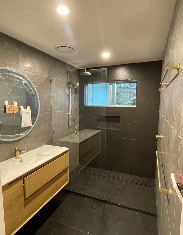 Modern bathroom with dark gray tiled walls and floor, a wooden vanity with a white sink and gold fixtures, a round mirror, a walk-in shower with a rainfall showerhead, a small window, and gold towel bars.