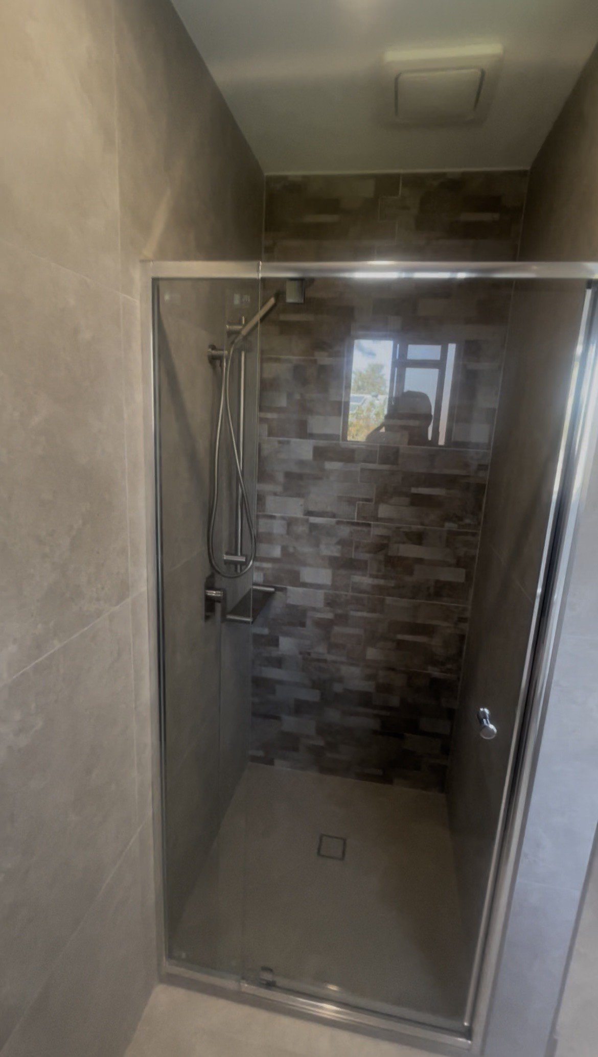 Modern shower with a glass door, beige and dark brown tiled walls, and a small window. Reflection of a person taking the photo is visible in the window.