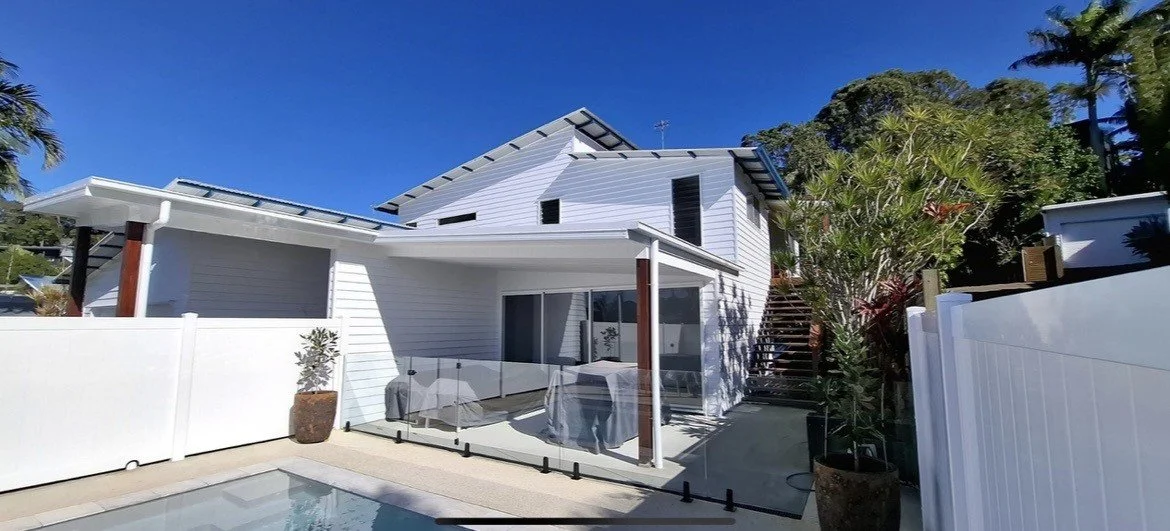 Modern white house with a small pool, outdoor patio area with barbecue grill, potted plants, and a staircase leading to the upper level, set against a bright blue sky.