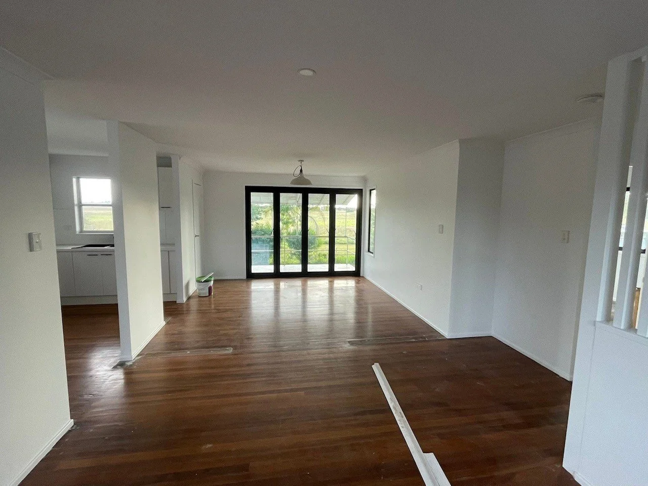 Empty living room with hardwood floors, white walls, large glass doors, and small windows, with some construction materials on the floor.