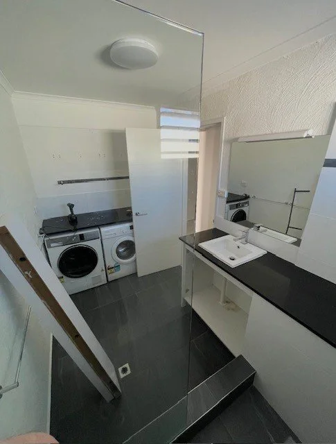 Small laundry room with a washer and dryer, a sink, a large mirror, and a door, with a glass partition.