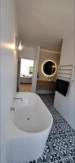 Modern bathroom with a white bathtub, patterned floor tiles, a towel rack with a black towel, a round backlit mirror, and a partial view of a living room with a television.