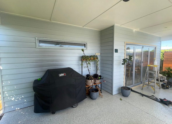 Patio area with a covered grill, potted plants, a ladder, and various gardening tools near a house with sliding glass doors.