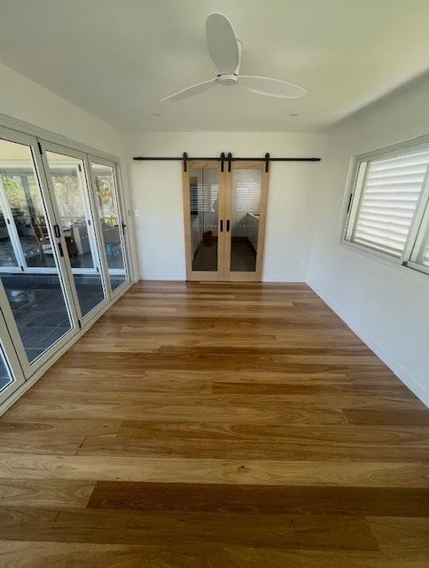 Empty sunroom with wooden floors, large sliding glass doors on the left, windows on the right, a sliding door at the back, and a ceiling fan.