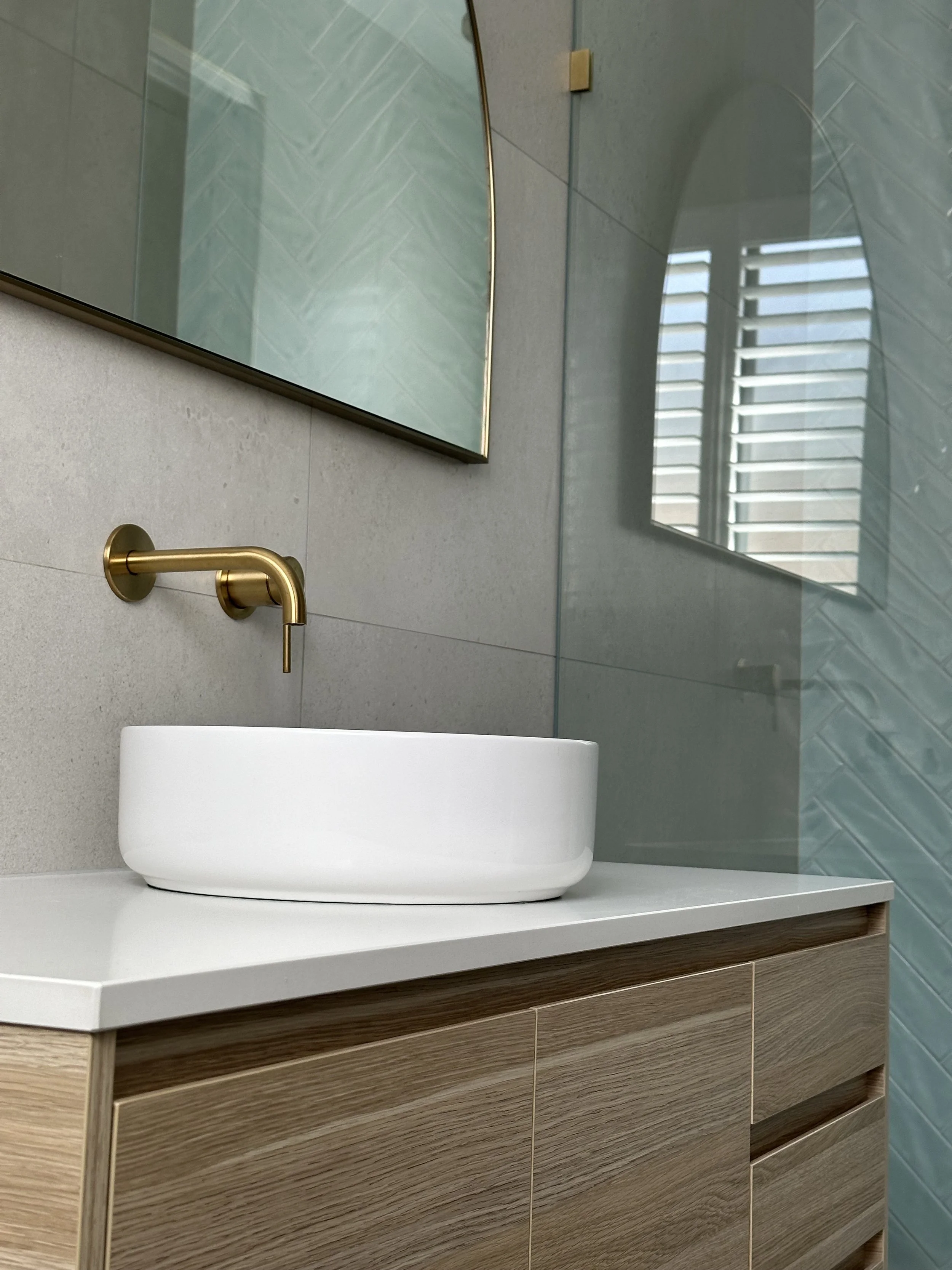 Bathroom with a vessel sink, wall-mounted gold faucet, wooden vanity, large mirror, textured teal wall tiles, and window with shutters.