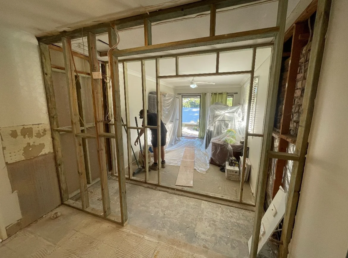 Room under renovation with exposed wooden framing and a person working, plastic sheets covering furniture, and a large window with green curtains.
