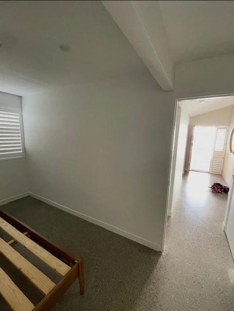 Empty room with white walls, a window with blinds, and an open doorway leading to a bright area with a door and window. Part of a wooden bed frame is visible in the foreground.