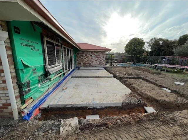 Construction site with a freshly poured concrete patio next to a house that is under renovation, with green building wrap and exposed brick, and a backyard with trees and a trampoline.