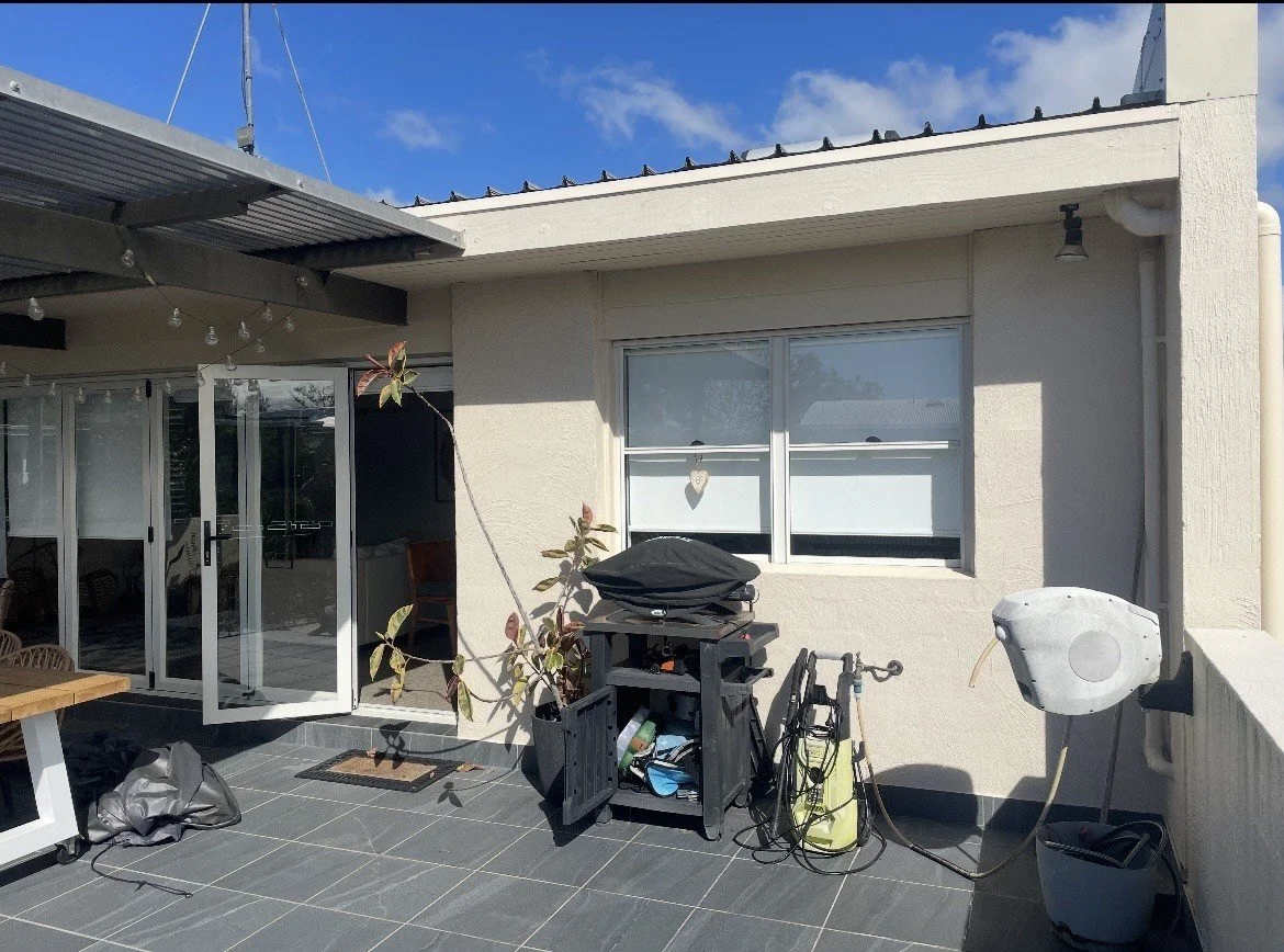 Outdoor patio with open glass doors, a plant, a black grill, a bicycle, and gardening equipment against a white wall with a window. A heart-shaped decoration hangs in the window, and a small wall-mounted light fixture is to the right.