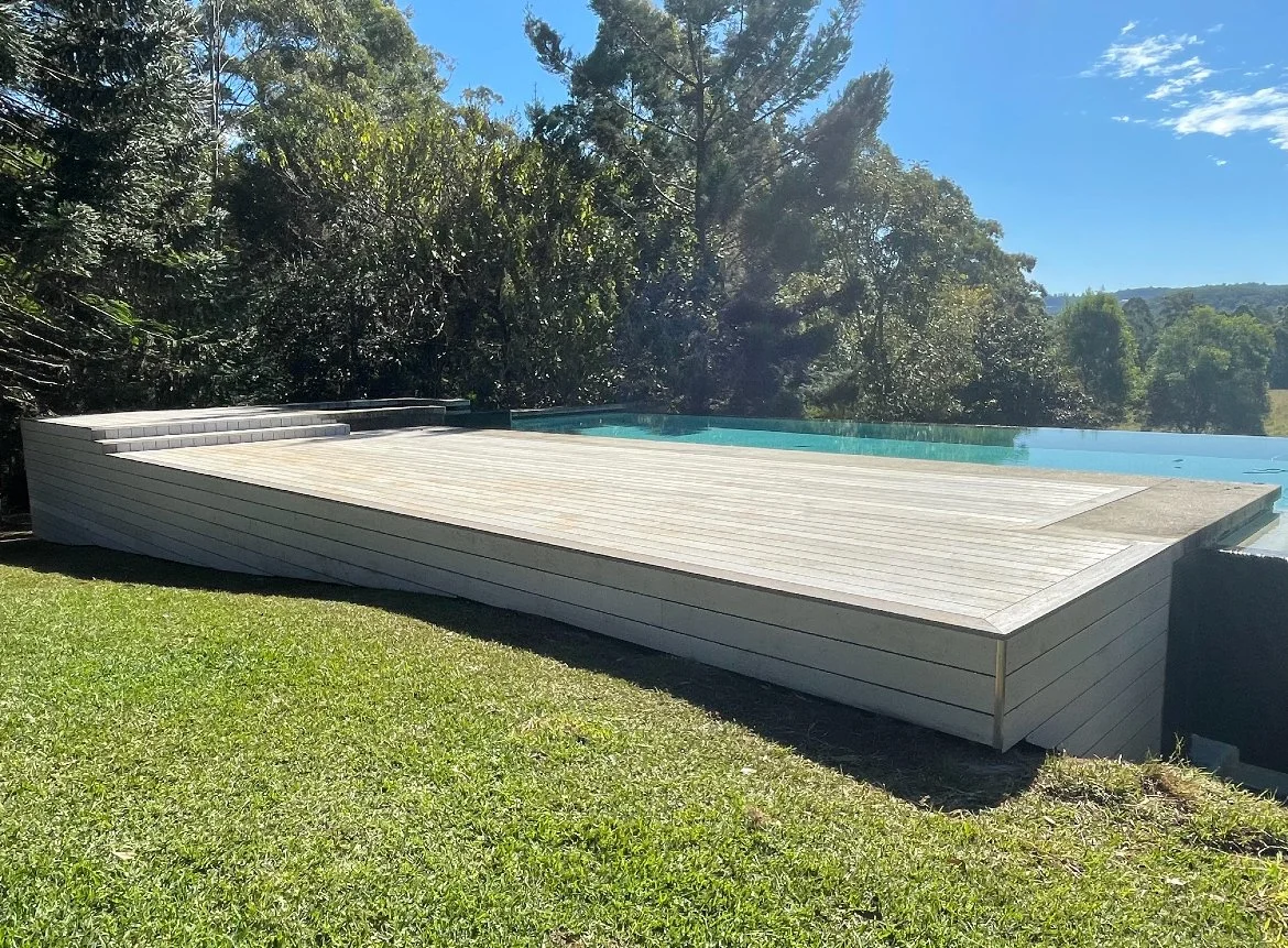 An outdoor pool with a wooden deck surrounding it, situated in a grassy backyard with trees and clear blue sky in the background.