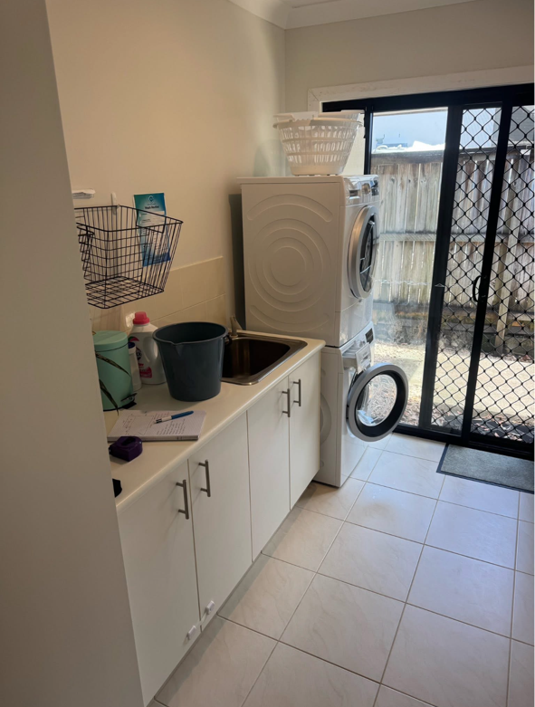Laundry room with white cabinets, a small sink, a washing machine and dryer stacked, a laundry basket on top, a black bucket near the sink, and natural light coming through sliding glass door with a fenced backyard outside.