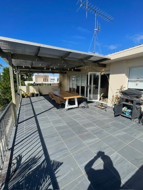 Outdoor rooftop patio with tiled flooring, seating area, barbecue grill, and an antenna on the roof under a blue sky.