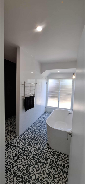 A modern bathroom with patterned floor tiles, a white bathtub, a towel rack with black towels, and a large window with blinds.