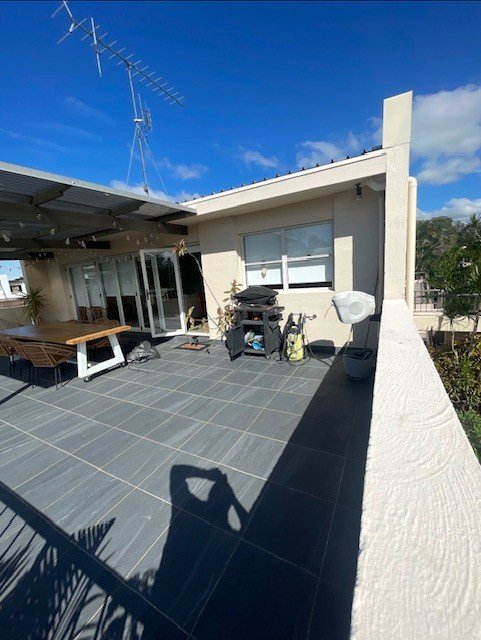 Rooftop patio with outdoor furniture, a barbecue grill, gardening supplies, and a glass sliding door, under a blue sky with clouds.