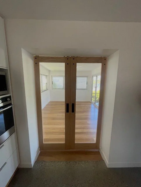View through wooden sliding glass doors into a bright, empty room with hardwood floors and multiple windows with blinds.