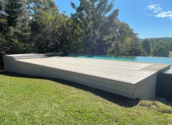 An outdoor above-ground swimming pool with a wooden deck in a backyard surrounded by greenery and trees, under a bright blue sky.