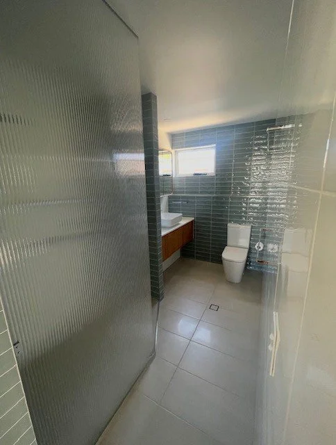 A modern bathroom with a toilet, a sink with a wooden cabinet, and a window with horizontal blinds. The walls are tiled with dark rectangular tiles, and the floor has large light gray tiles.