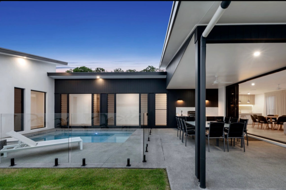 Modern house backyard with swimming pool, sun lounge, outdoor dining area with black chairs, glass boundary, and sliding glass doors leading inside, during evening with lights on.
