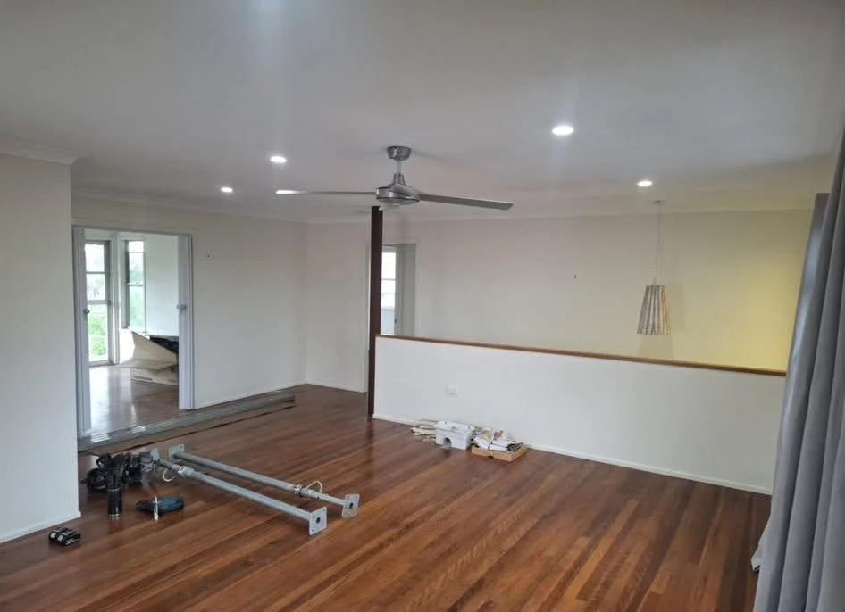 Empty living room with hardwood floors, white walls, a ceiling fan, and recessed lighting. There are some construction or installation items on the floor and an open doorway leading to another room with large windows and a door to a balcony.