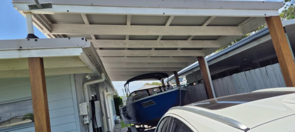 A covered carport with a boat stored underneath it, positioned beside a house and a vehicle parked nearby.