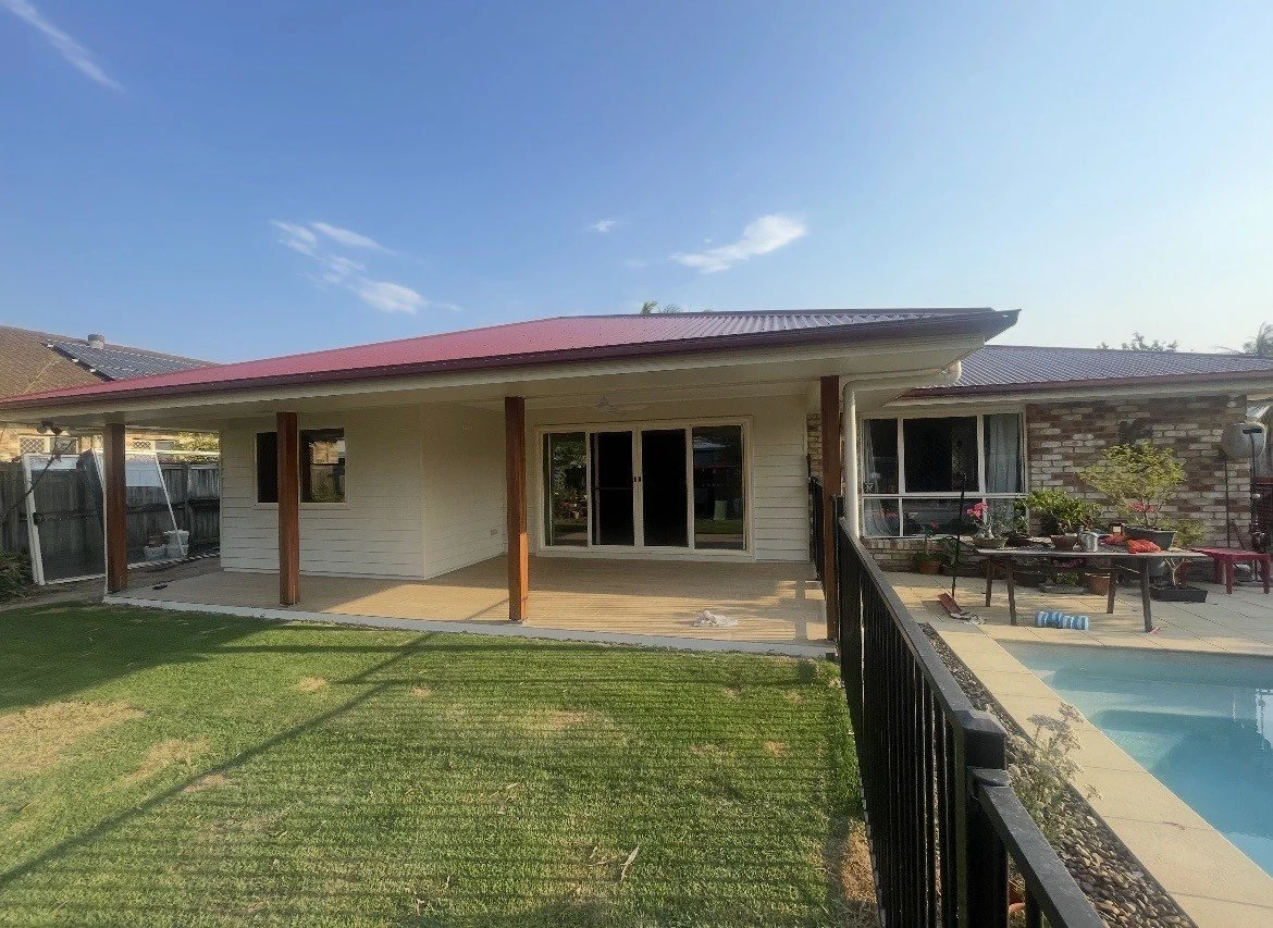 Backyard view of a house with a covered patio, sliding glass doors, a swimming pool, and a fenced lawn, during daytime.