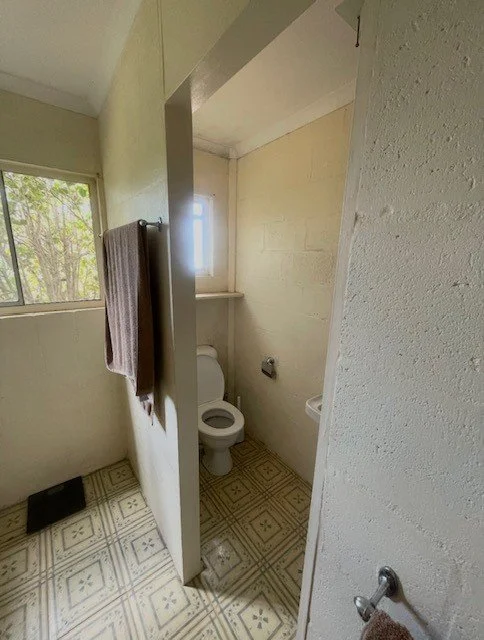A small bathroom with a toilet, a small window, and part of a sink. A brown towel is hanging on a towel rack on the wall. The floor has patterned tiles, and there is a small black rectangular object near the window.