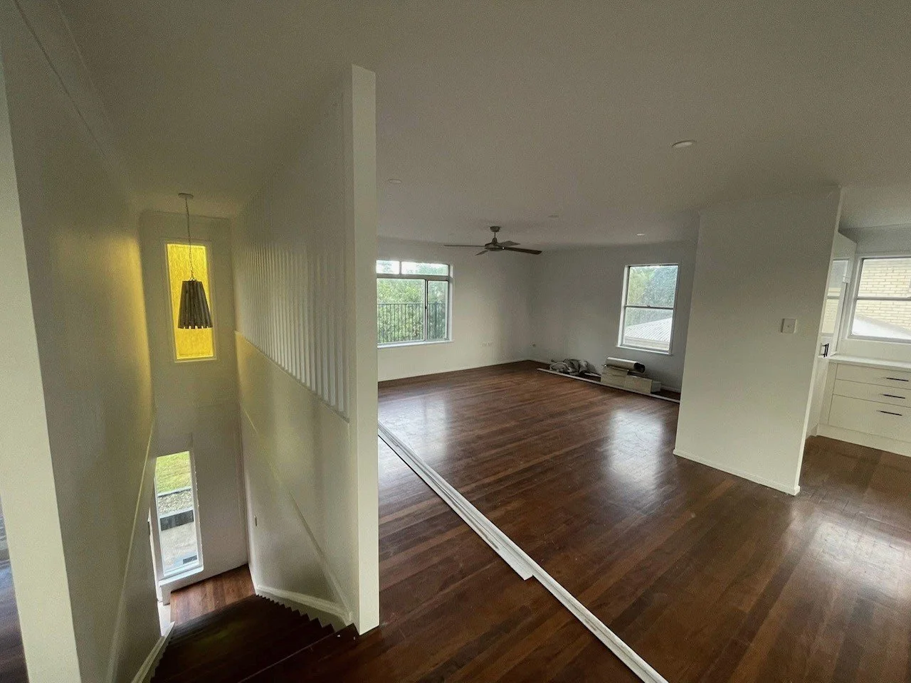Empty living room with hardwood floors, large windows, ceiling fan, and white walls. Part of a staircase with a yellow-covered hanging light nearby.