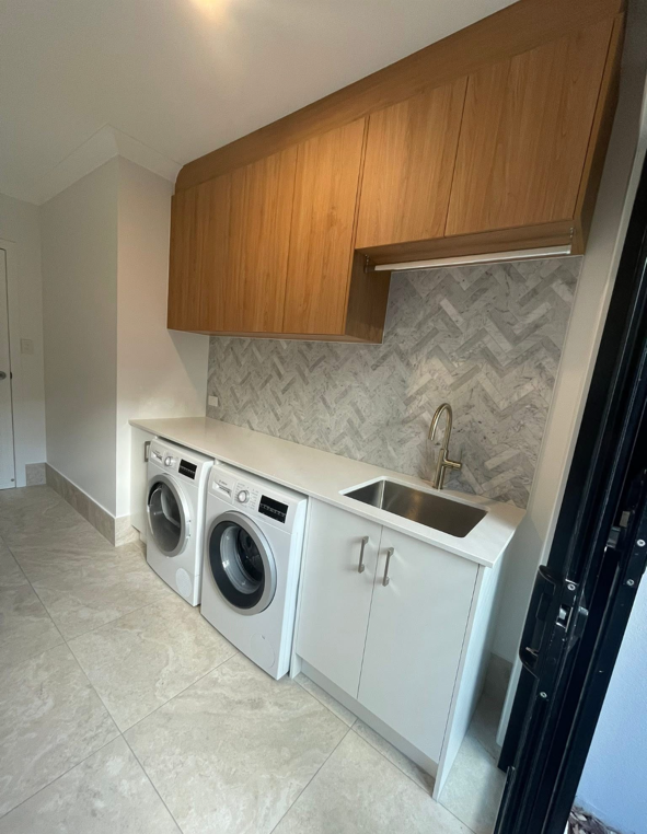 Laundry room with white washing machine and dryer, white counter with sink, brown cabinets above, gray patterned backsplash, and light-colored tiled floor.