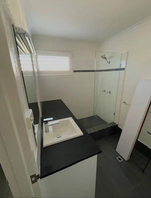 Bathroom with a black countertop, white sink, small mirror, shower with glass door, and a window with frosted glass.